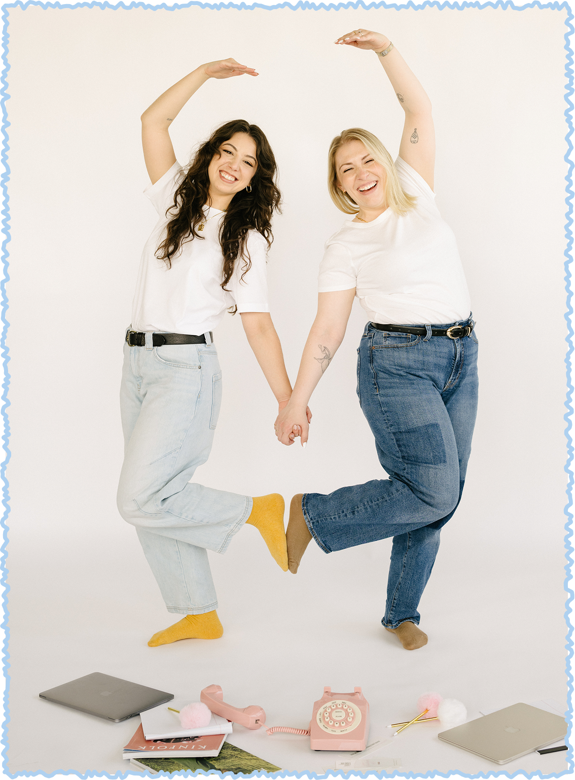 Two women holding hands, wearing white t-shirts and jeans, smiling, with one wearing yellow socks and the other wearing brown socks, standing on one leg each, with a vintage pink rotary phone, laptops, a notebook, and a magazine on the floor in front of them.