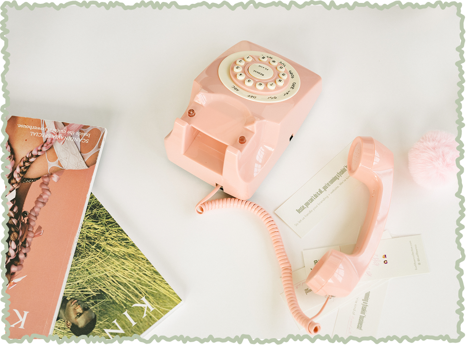 Pink rotary dial telephone with coiled cord, a pink pom-pom and magazines on a white surface.