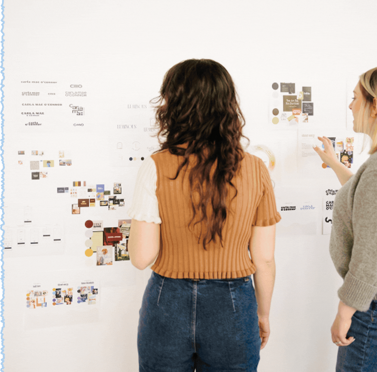 Two women discussing a large whiteboard filled with various images, texts, and graphics in an office setting.
