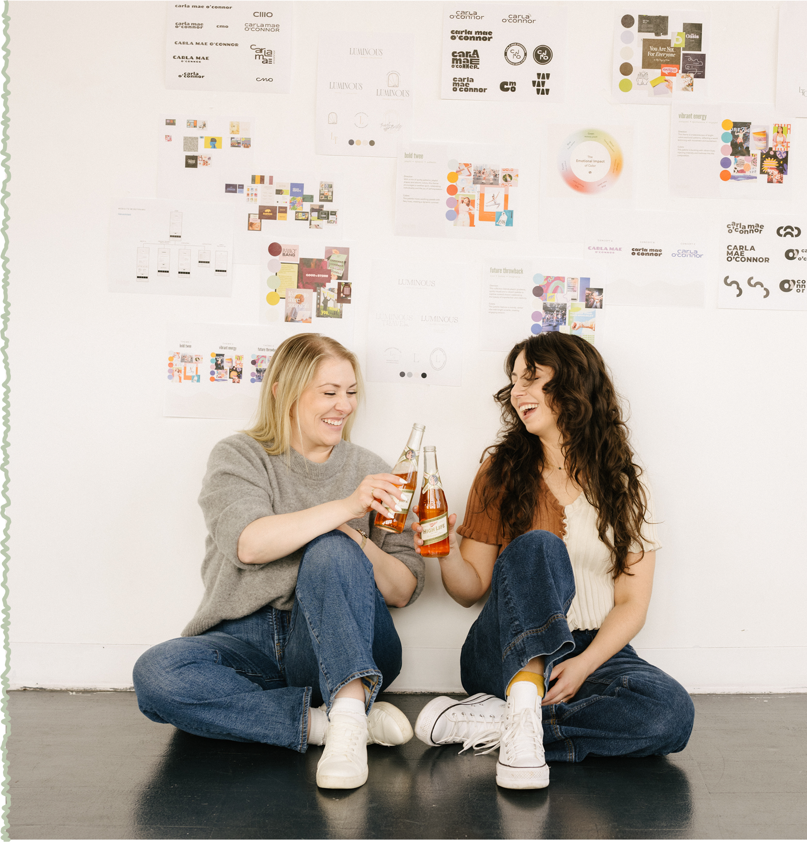 Two women sitting on the floor, smiling and clinking bottles of beer together, with a white wall and various colorful posters behind them.