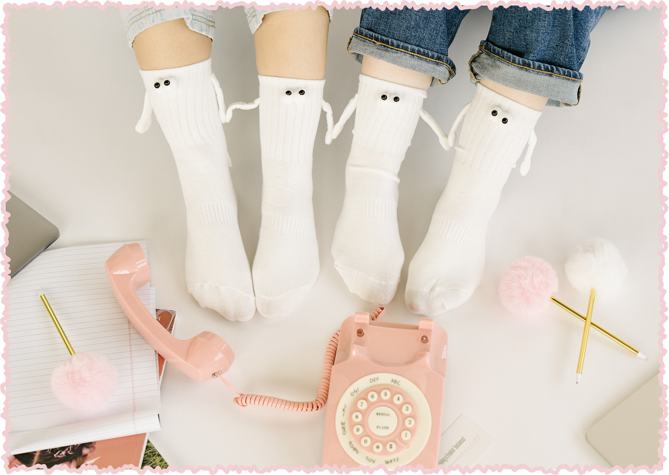 Two people sitting together wearing white socks with small black eyes and sock arms, holding hands. There is a pink vintage rotary telephone, two pink fluffy pens, a notebook, and a closed silver laptop on a white floor.