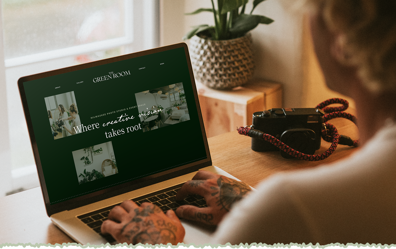 A person with tattoos on their hands using a laptop with a website titled "The Green Room," featuring interior design images. There is a camera with a red and black strap on the wooden table next to them, and a potted plant in the background.