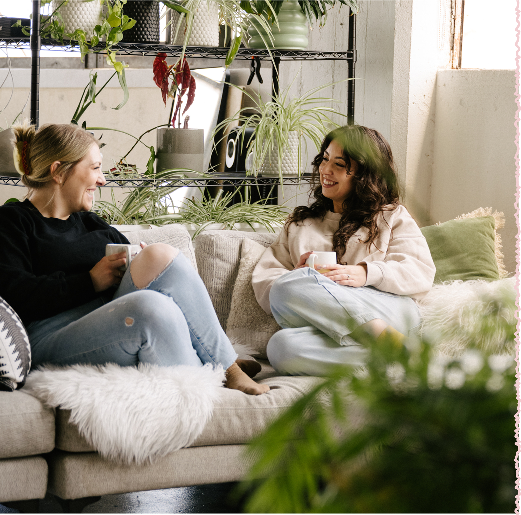 Two women sitting on a beige sofa, smiling and holding mugs, in a cozy, plant-filled living room with natural light.