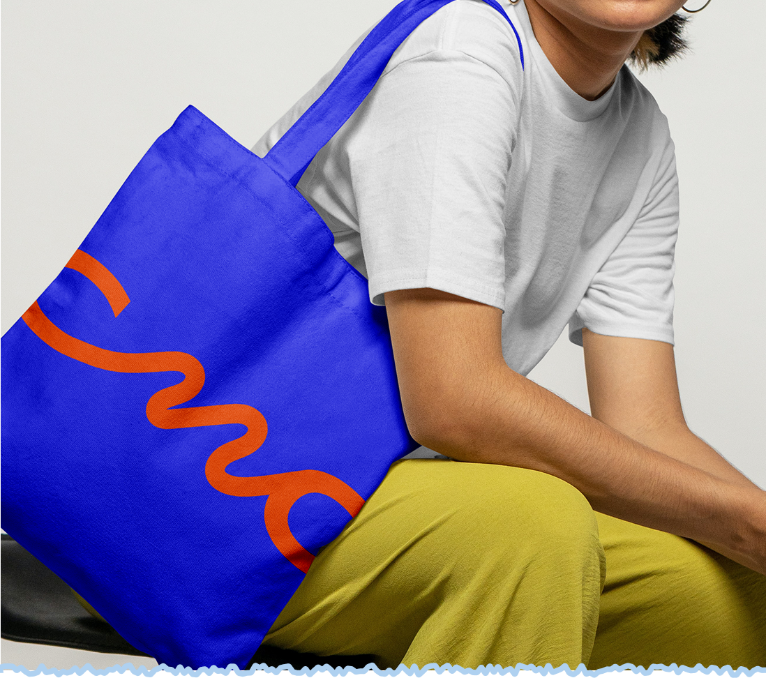A person sitting on a black chair with a blue tote bag with orange writing saying CMO (Carla Mae O'Connor), wearing a white t-shirt and yellow pants.