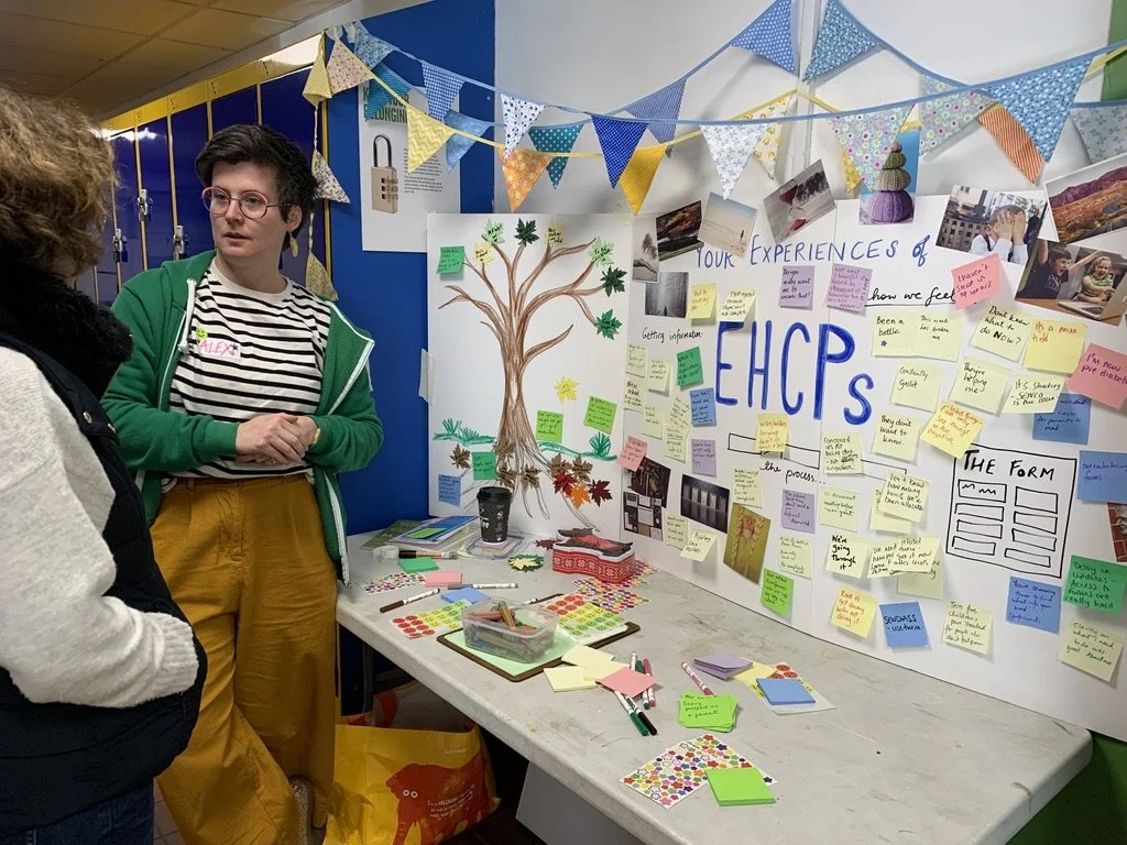 Alex listening to a parent at a Family Fun Day, standing beside a visual board covered with drawings, photos and notes about families’ experiences of SEND and EHCPs.