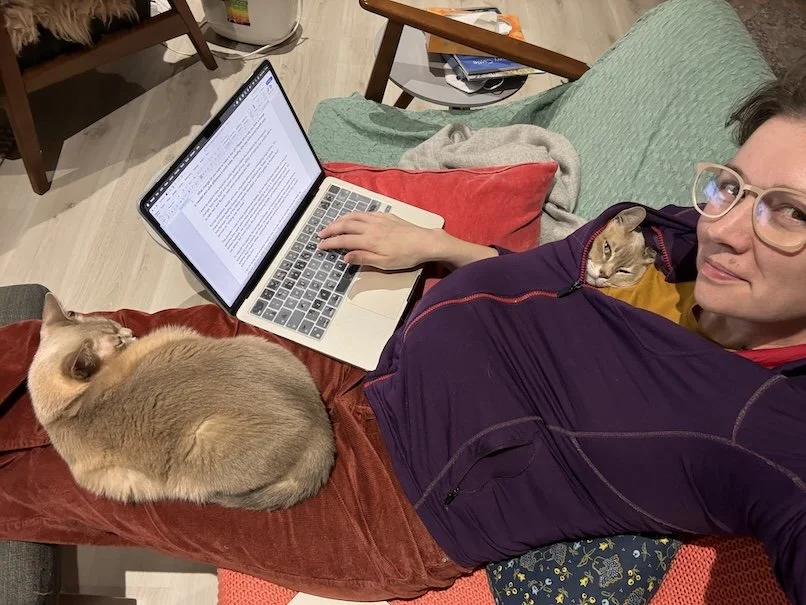 A woman sitting on a sofa working on a laptop, with two cats resting close by in a shared living space.