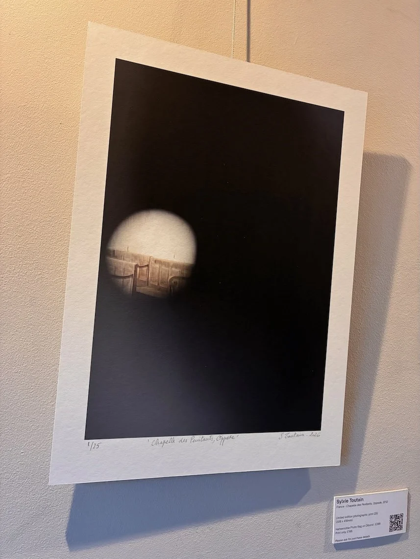 A photographic print depicting a church interior glimpsed through a small circular opening, surrounded by darkness, highlighting how much of the scene remains unseen.