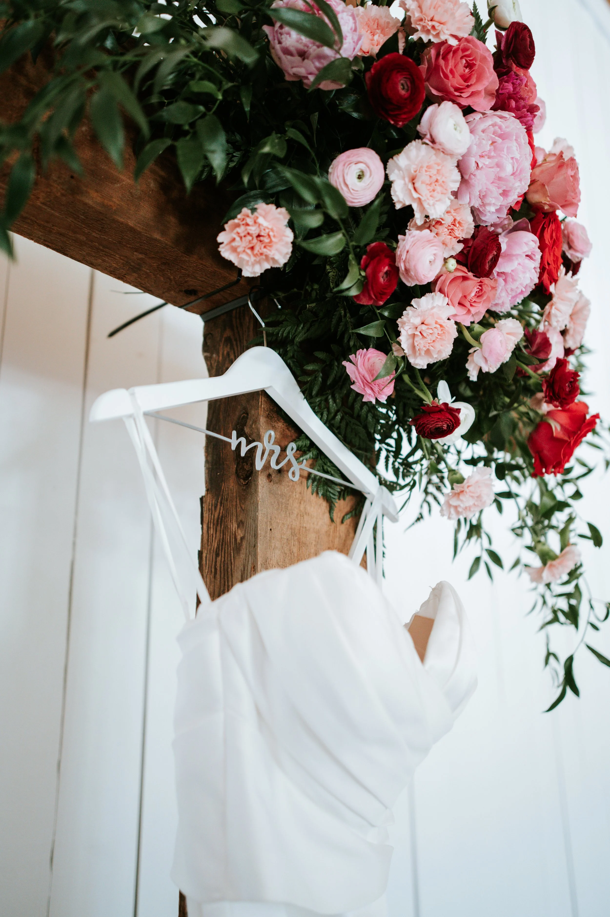 various pink colored florals and greneery hung arangemenr on a wooden cross with the wedding dress hanging below it
