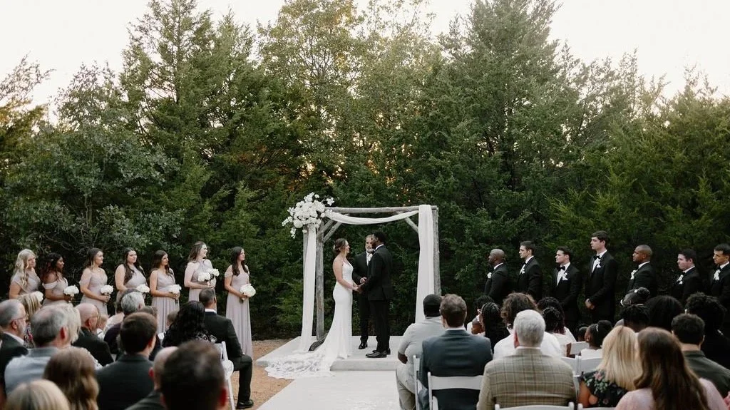 Outdoor wedding ceremony with bride and groom exchanging vows under an archway, surrounded by bridesmaids and groomsmen, and seated guests watching.