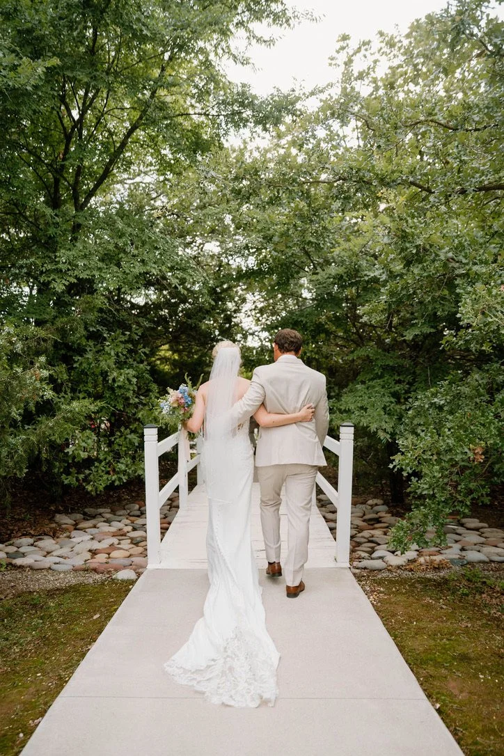 Bride and groom walking arm in arm on a small white bridge surrounded by green trees, with the bride holding a bouquet of flowers.