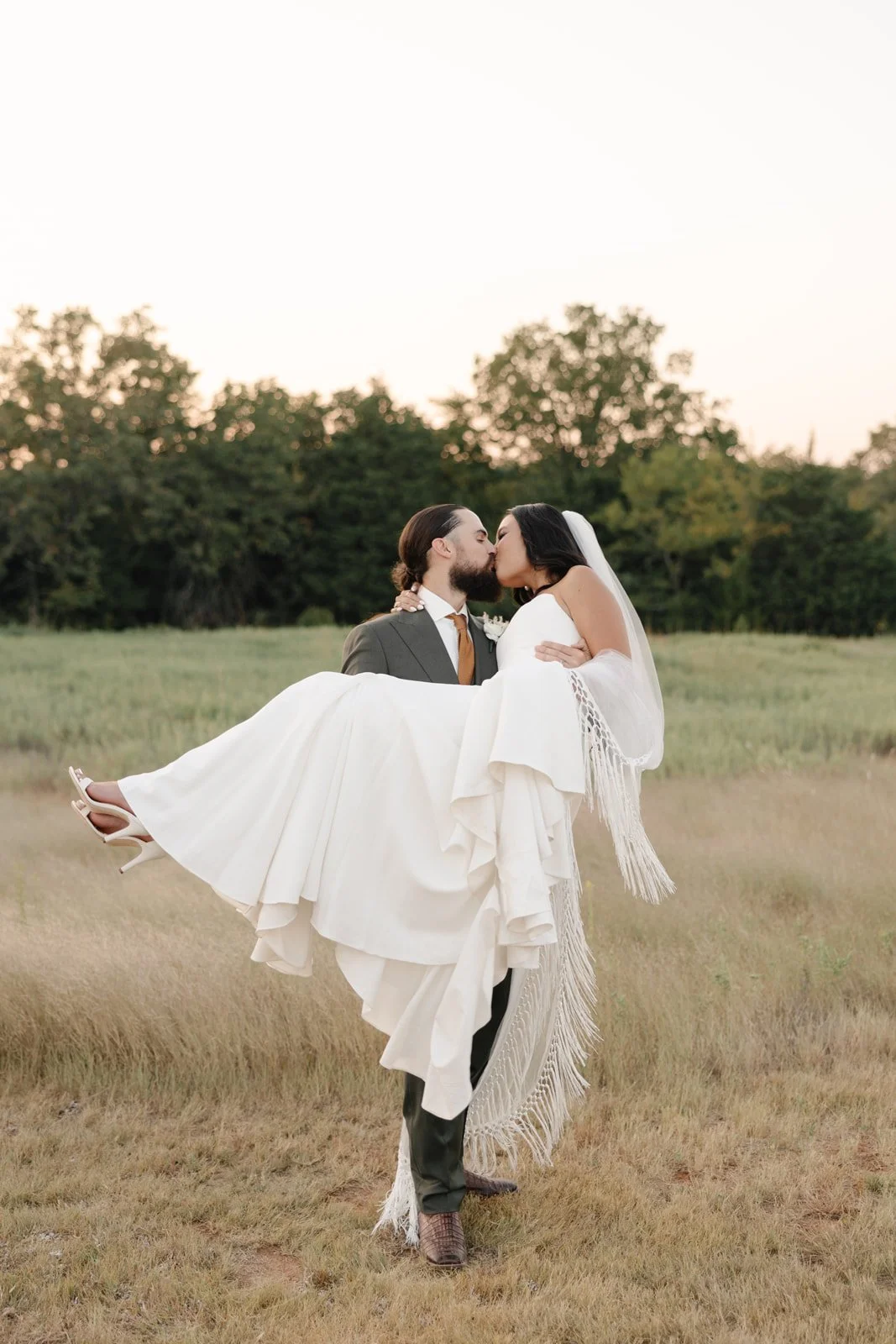 A newlywed couple kiss in an open field at sunset, with the groom carrying the bride in his arms. The bride is wearing a white wedding dress and veil, while the groom is dressed in a suit and tie. The background features trees and a softly lit sky.