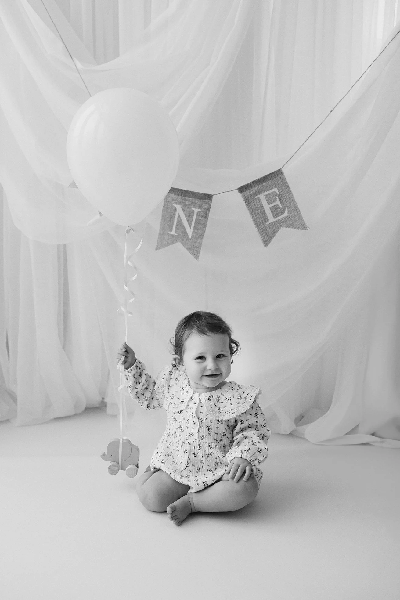 A smiling toddler girl sitting on the floor holding a balloon with a string, decorated with a banner that spells 'NE' in the background, set against a backdrop of draped fabric.