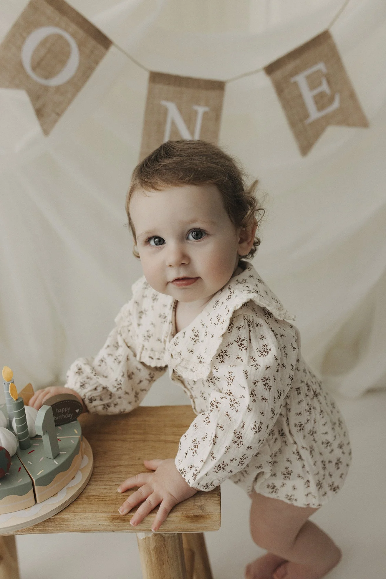 A young girl with curly brown hair, wearing a patterned cream-colored dress, is kneeling at a wooden table with a birthday cake in front of her. Behind her, there is a beige 'ONE' banner hanging on the wall.