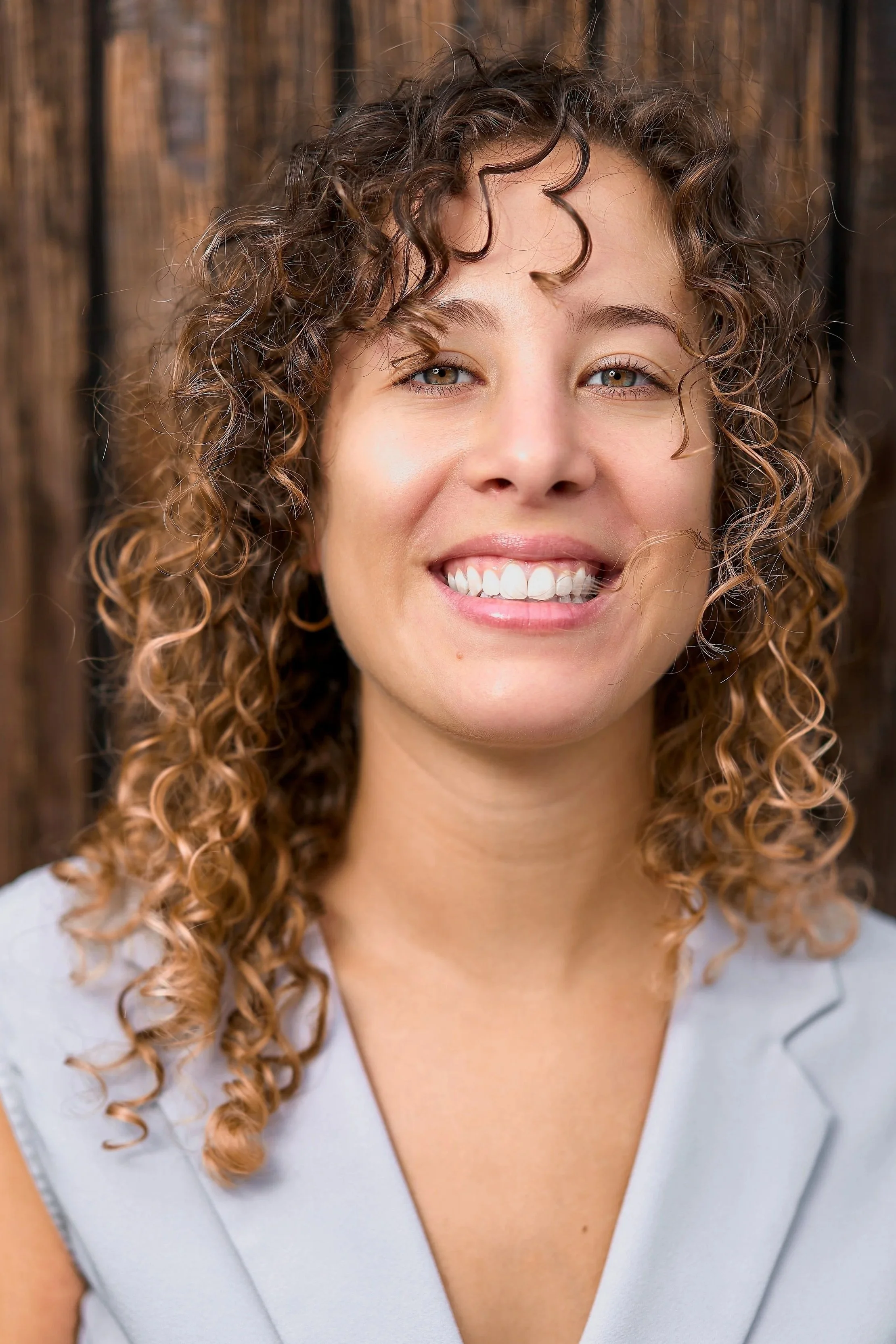 Close-up of a young woman with curly brown hair, smiling, wearing a light gray top, and standing in front of a wooden background.