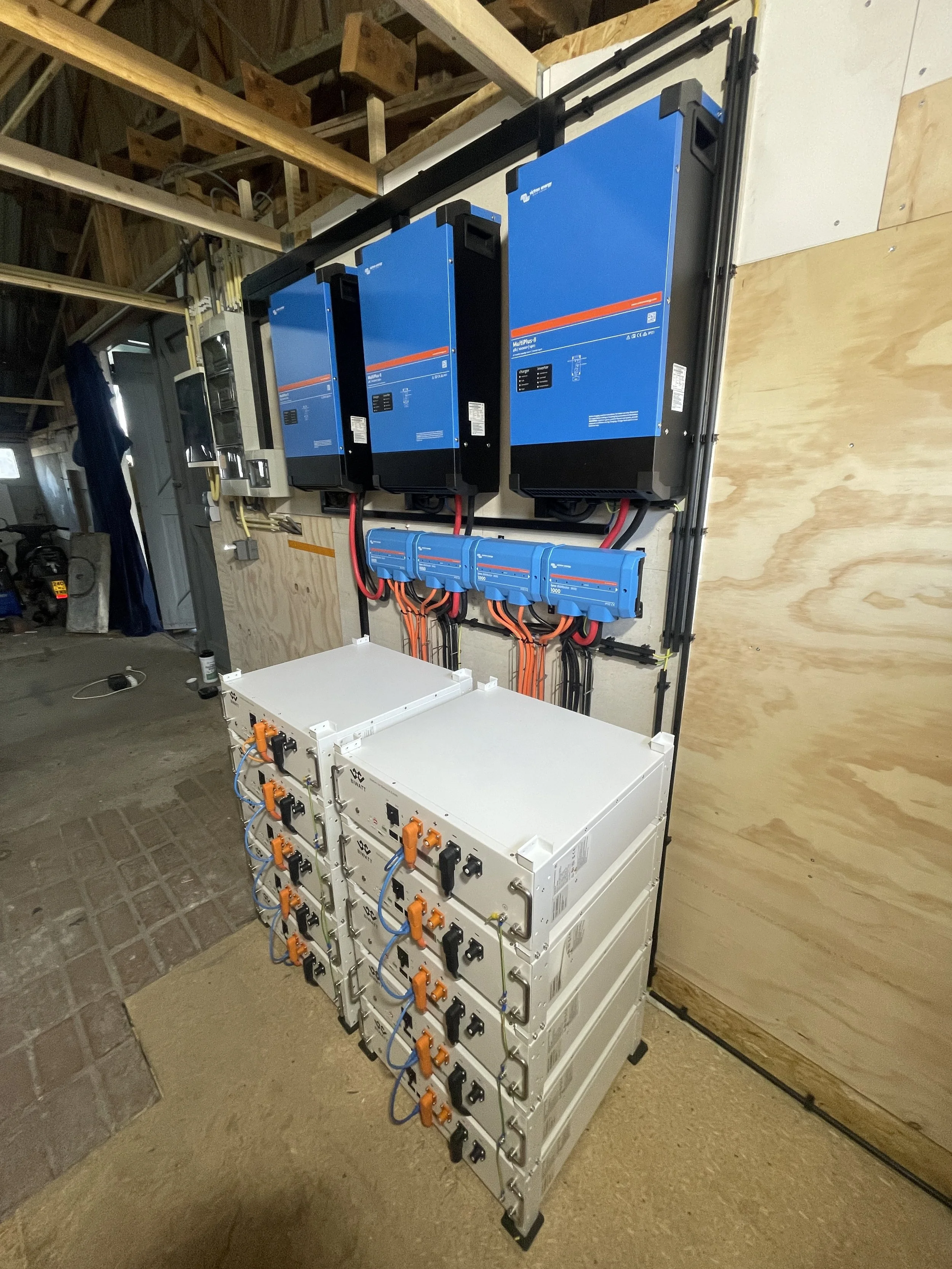 Elektrisch gemodificeerde installatie met drie grote blauwe omvormers, blauwe scheidingsblokken, en meerdere witte kalfom onderdelen met orange en zwarte connectors, geïnstalleerd op een houten muur.