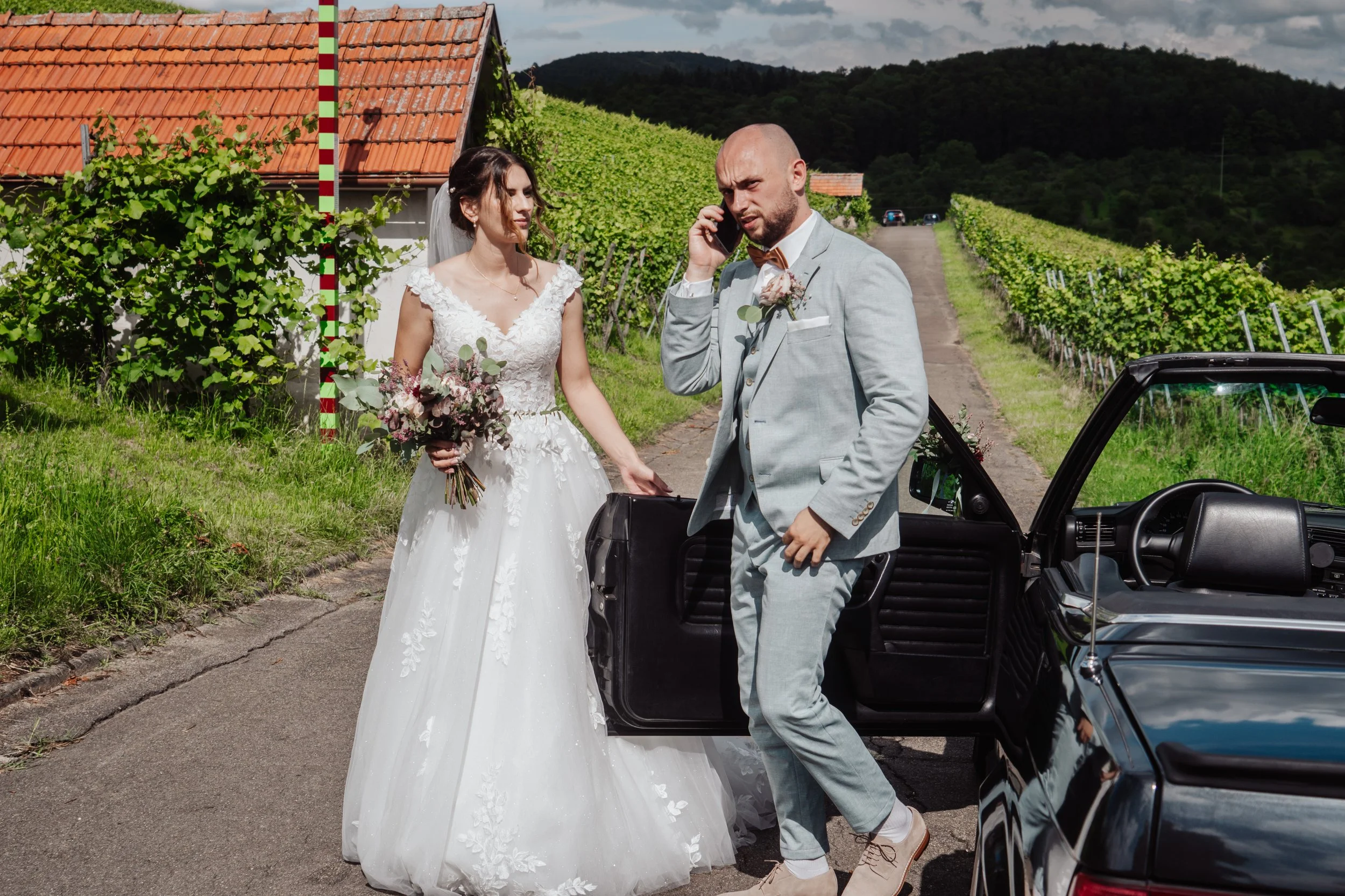 Braut in weißem Kleid und Bräutigam in Grau, die mit einem Auto unterwegs sind, in einer ländlichen Gegend mit Weinreben im Hintergrund, bei Sonnenlicht.