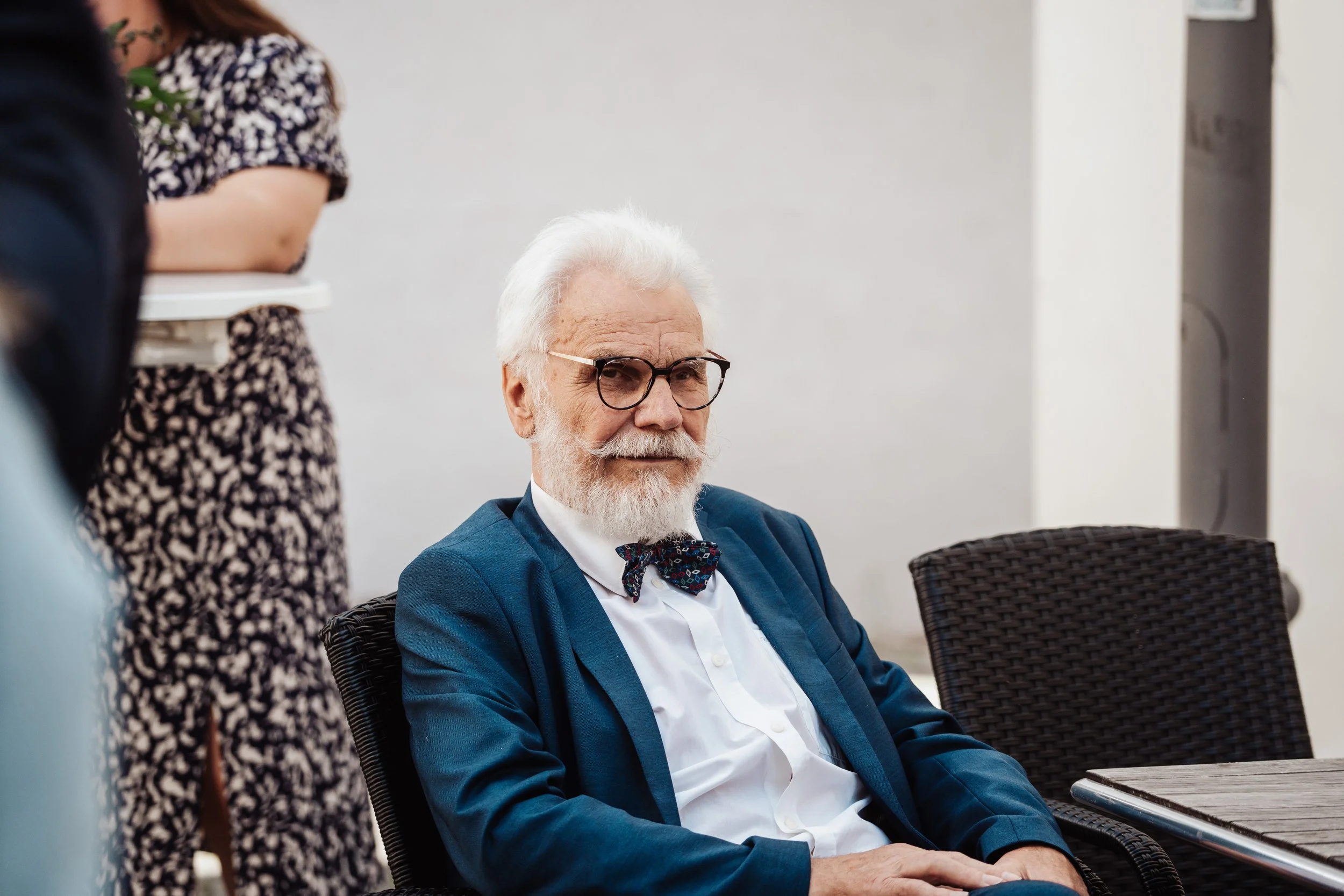 Ein älterer Mann mit weißem Haar und Brille, trägt einen blauen Anzug und Fliege, sitzt an einem Tisch.