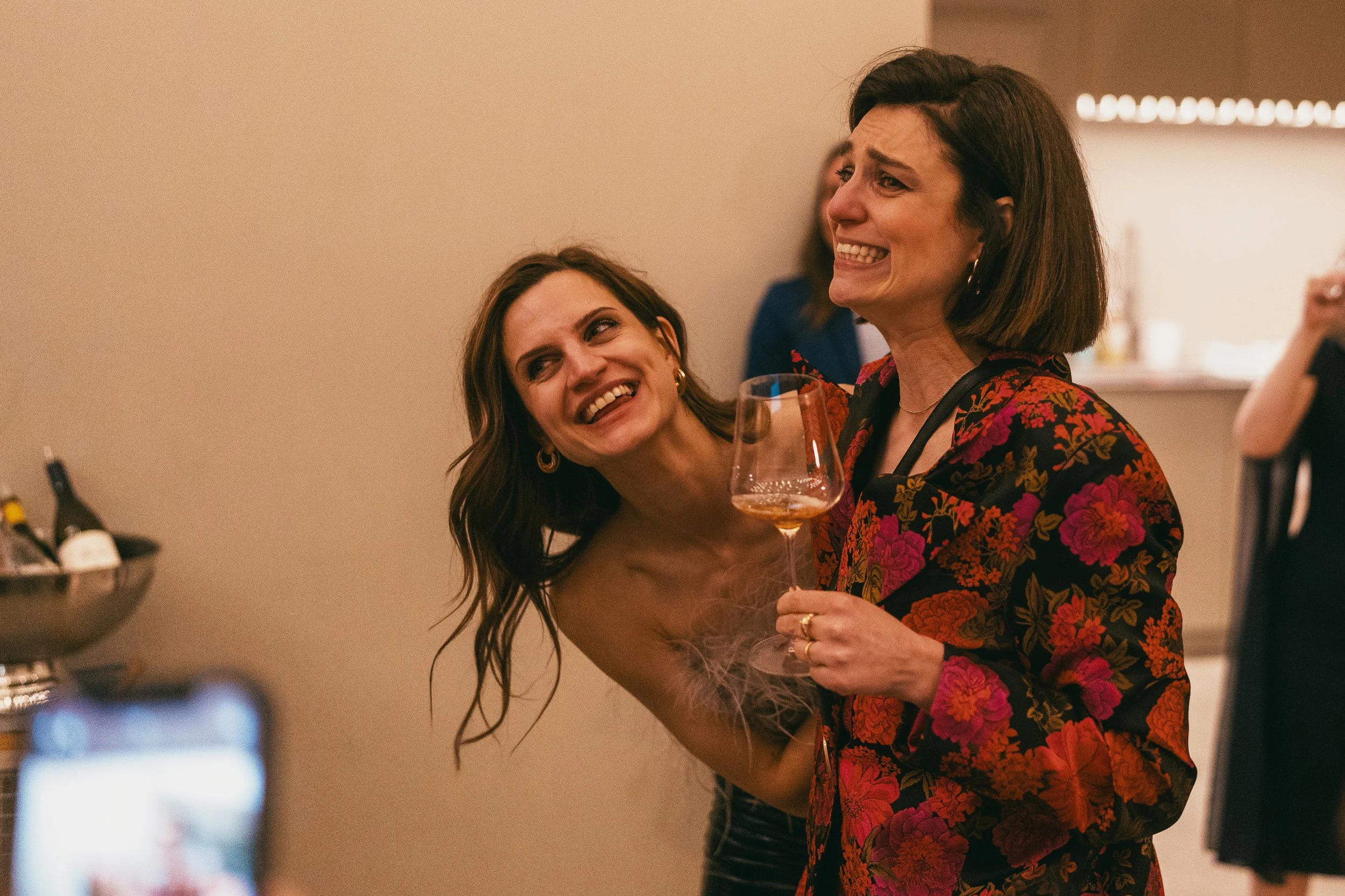 Zwei Frauen lachen und haben Spaß auf einer Party, eine hält ein Weinglas, die andere steht hinter ihr, im Hintergrund unscharfe Beleuchtung und andere Personen. Hochzeitsfotograf Stuttgart | Fotograf | Filmproduktion Stuttgart | Videograf | Contentc