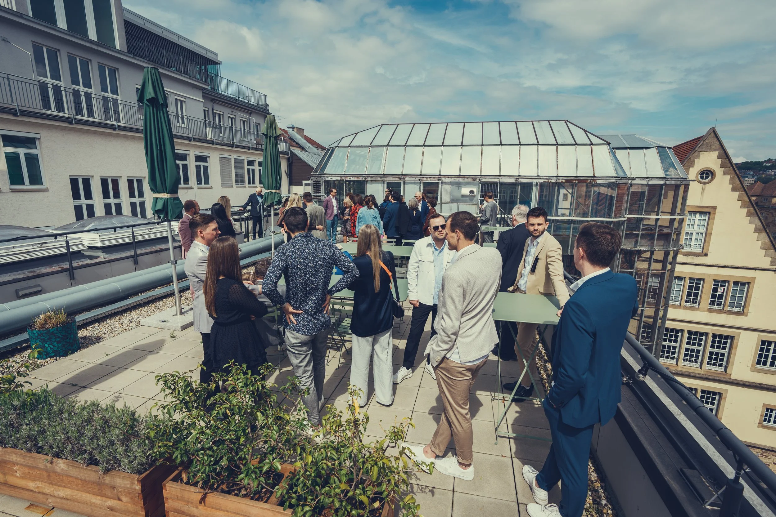 Menschen stehen auf einer Dachterrasse im Gespräch, im Hintergrund ein gewächshausartiges Glasgebäude und begrüntes Umfeld, sonniger Himmel.