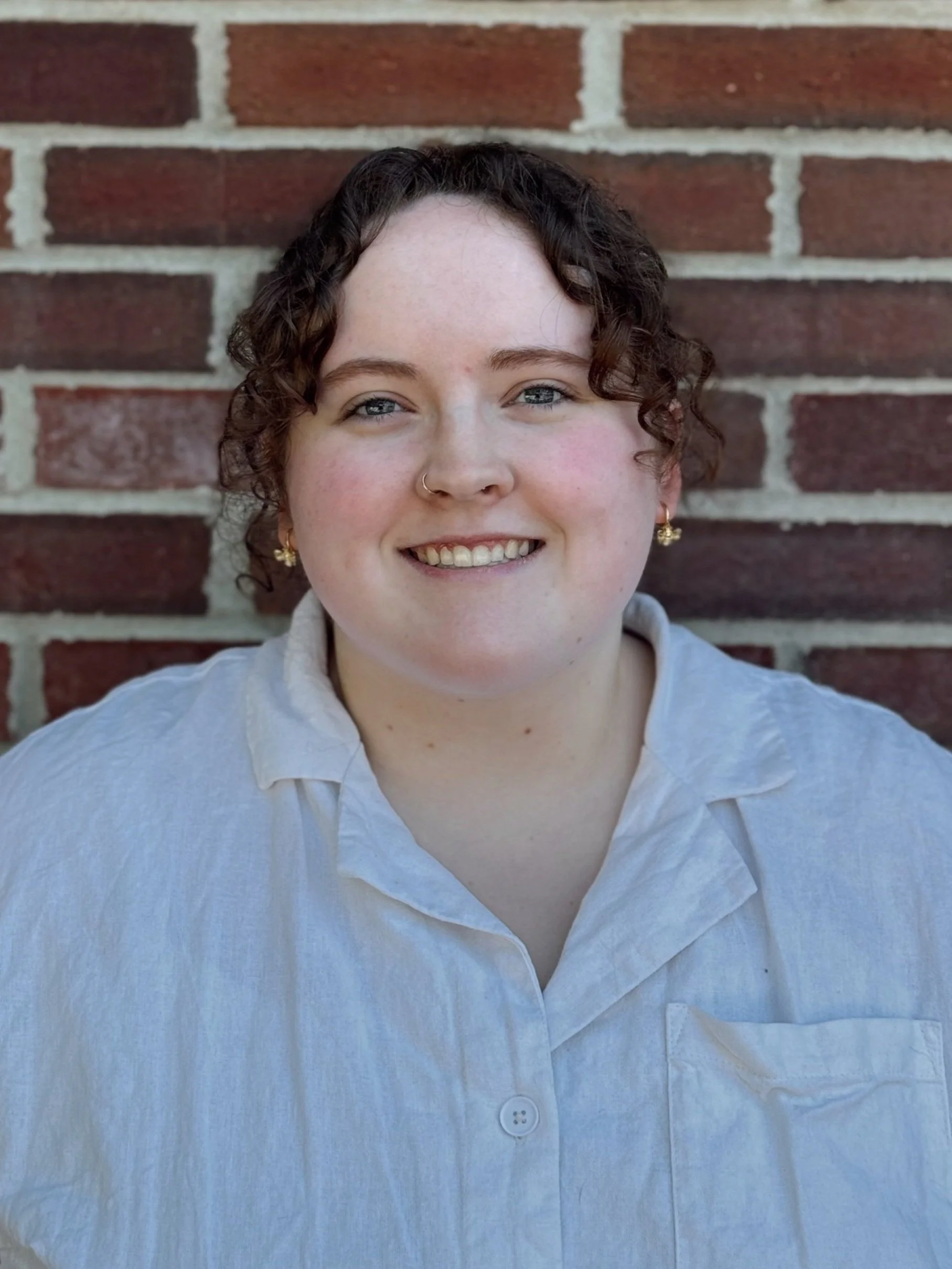 A woman with curly brown hair and blue eyes smiling, wearing a white button-up shirt, standing in front of a brick wall.