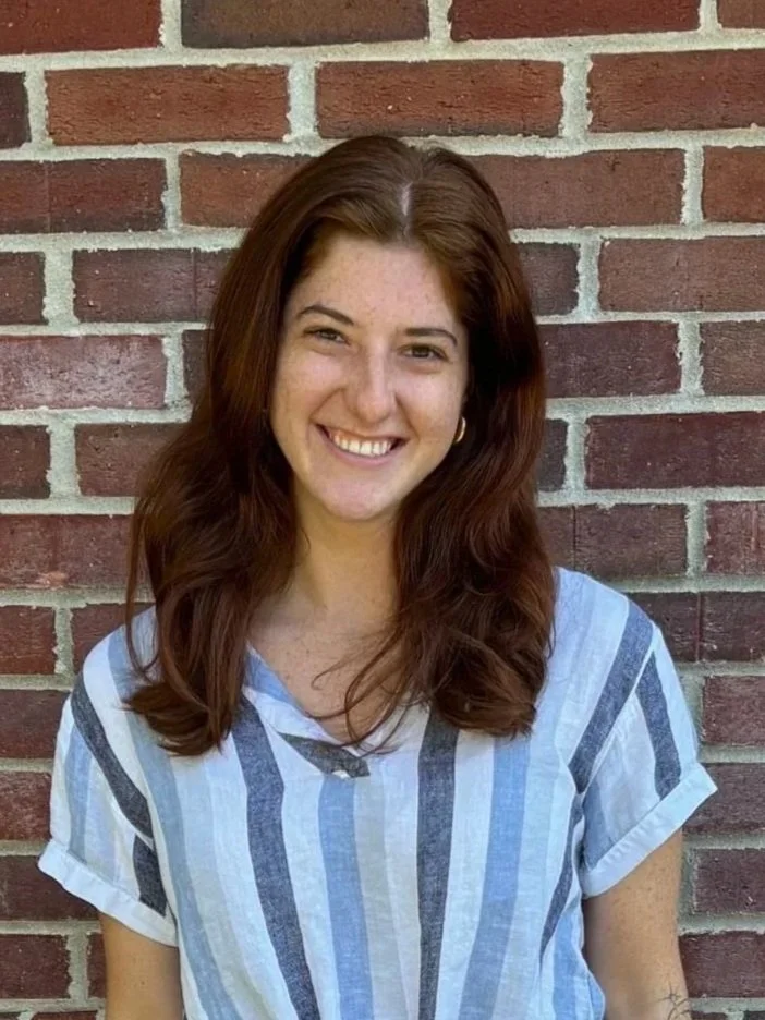 A young woman with long, wavy brown hair smiling, standing in front of a brick wall, wearing a striped short-sleeve shirt.