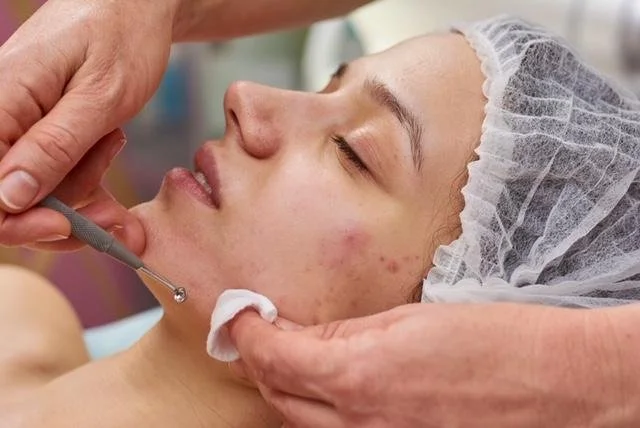 A woman lying down with her eyes closed, wearing a hair cap, receiving a skincare treatment with a dropper and cotton pad.