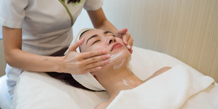 A woman receiving a facial treatment with a professional applying a cream on her face, lying on a spa bed.