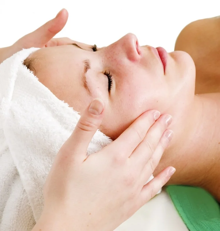 Person receiving facial massage while lying down with eyes closed, wrapped in a towel.