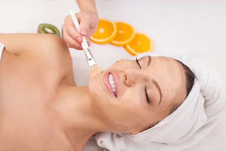 A woman receiving a facial treatment with a brush on her face, wearing a towel wrapped around her head, with slices of orange and kiwi fruit on the table