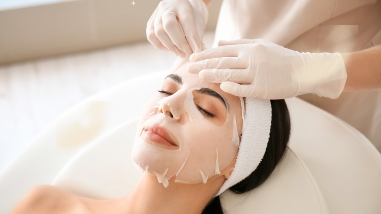 A woman receiving a facial treatment while lying on a spa bed with her eyes closed, a white headband keeping her hair back, and a beautician applying a facial mask or peel.
