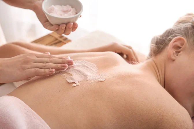 A person receiving a Himalayan salt massage, with pink Himalayan salt being spread on their back, while another person holds a bowl of pink Himalayan salt.