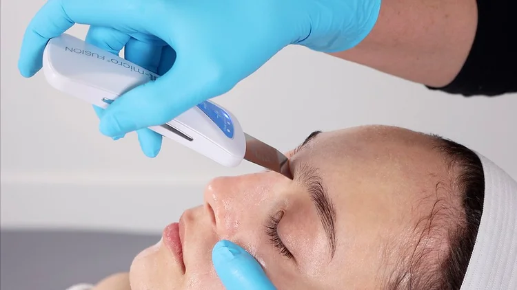 A person is receiving a cosmetic or medical treatment in a clinical setting, with a gloved hand holding an ultrasonic device on their forehead while their face is relaxed.