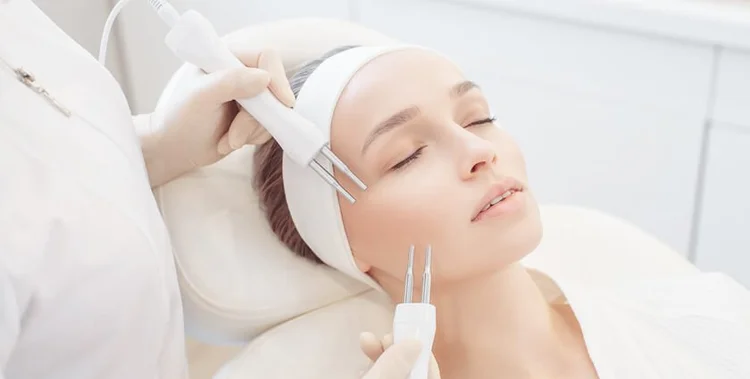 A woman receives a facial treatment with a handheld device in a clinic; she is lying down with eyes closed, wearing a headband.