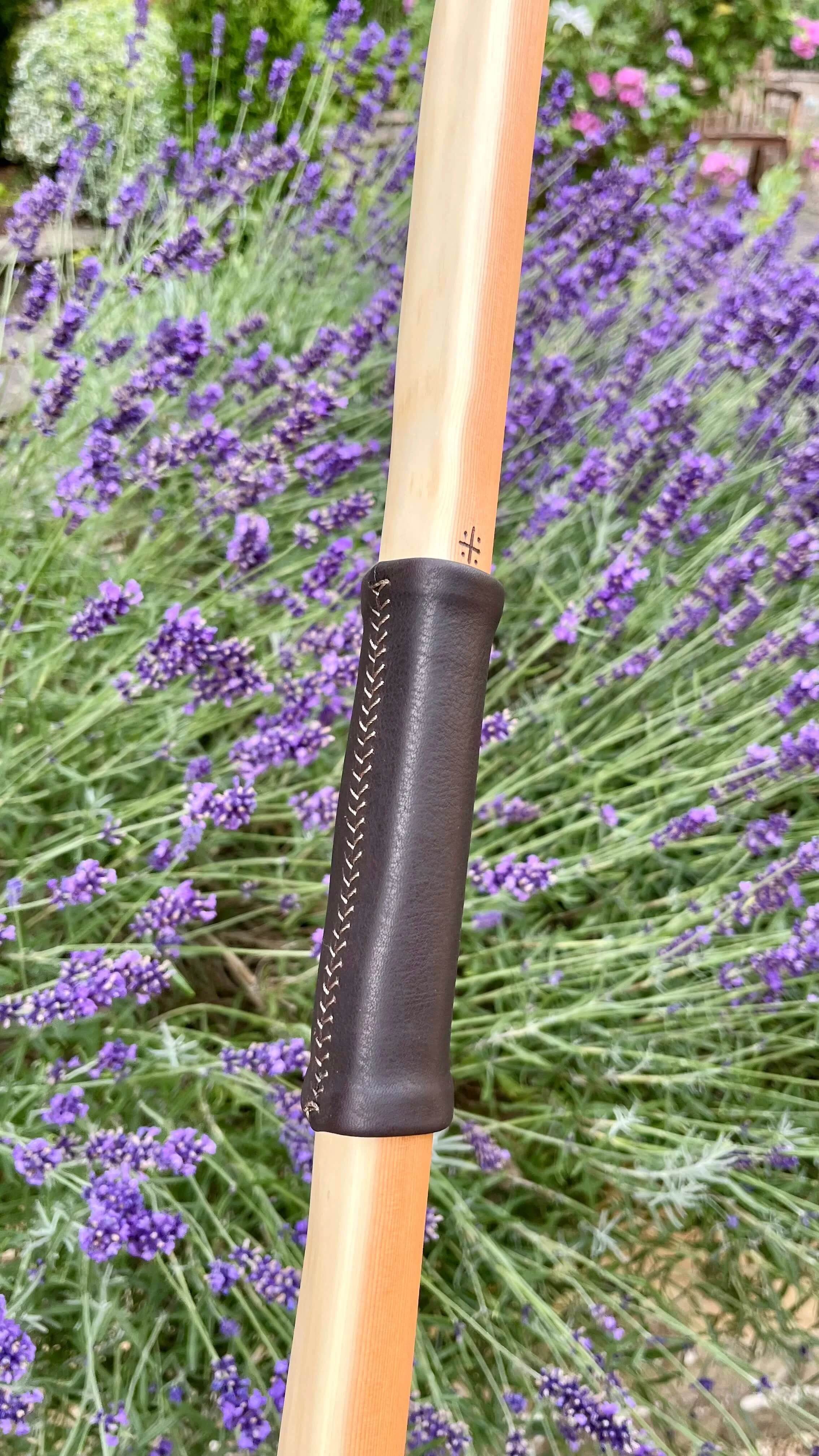 Close-up of a dark brown, hand-stitched leather handle grip on an archery bow against purple lavender flowers.