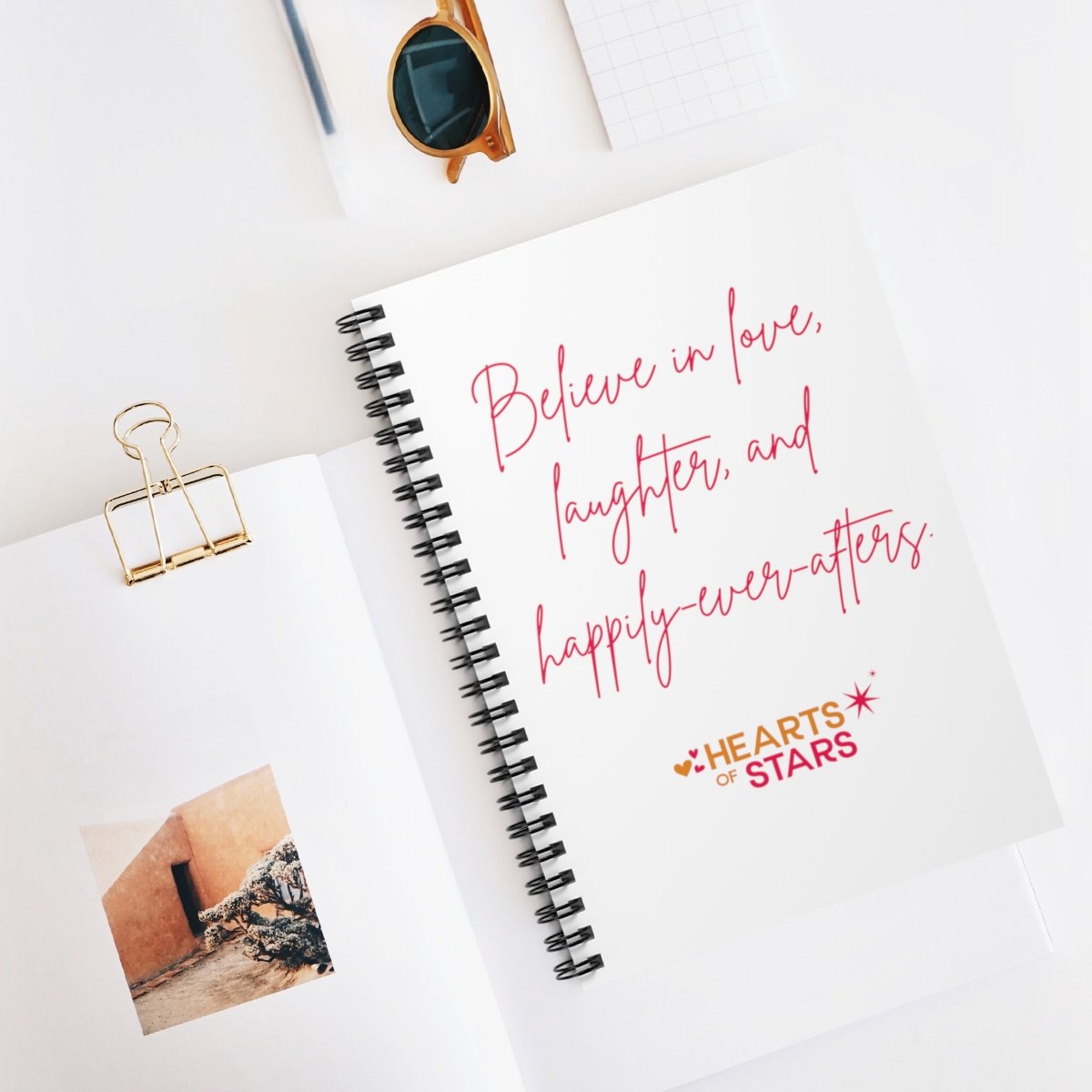 Flatlay of stationery including spiral notebook with "Believe in love, laughter, and happily-ever-afters" text, sunglasses, paperclip, and photo print.
