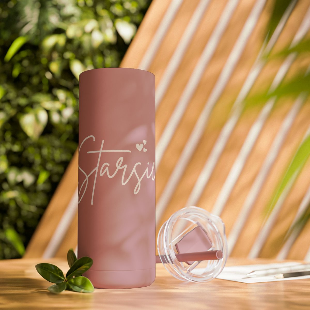 Pink insulated tumbler with script text and lid on wooden table, greenery background.