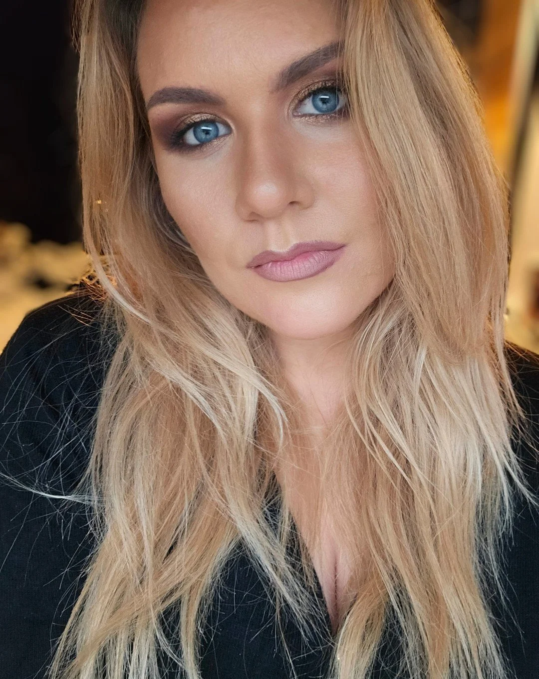 A woman with long, blonde hair and blue eyes looking at the camera, wearing makeup and a black top.