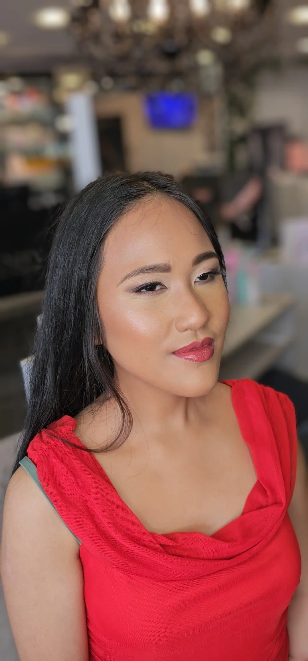 A woman with long black hair and makeup wearing a red dress, posing in an indoor setting with a blurred background.