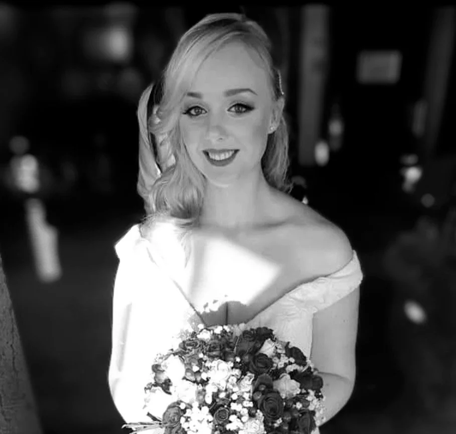 A woman with wavy blonde hair in an off-shoulder dress holding a bouquet of flowers, smiling, in a black-and-white photo.