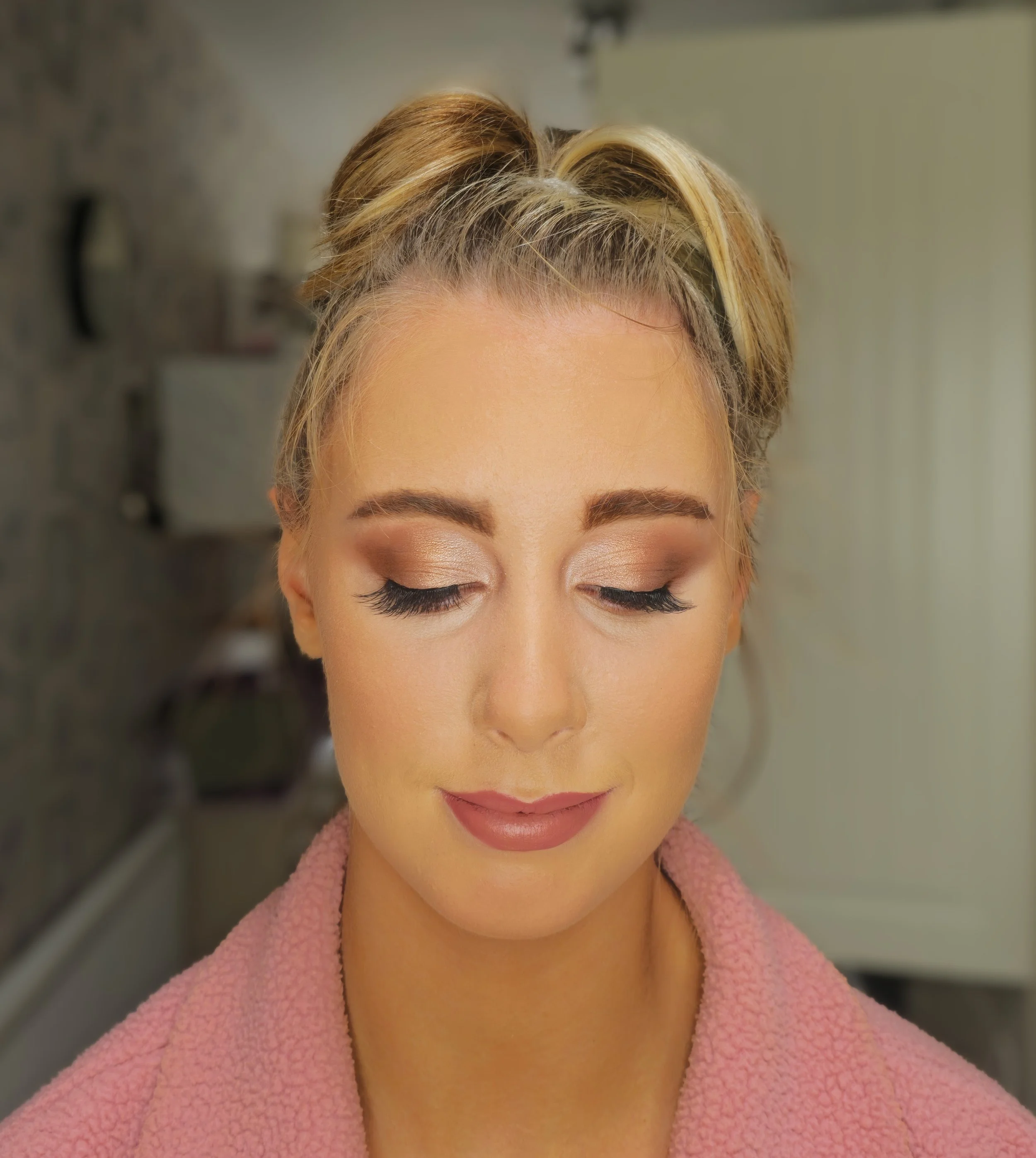 Close-up of a woman with styled blonde hair, closed eyes, and natural makeup, wearing a pink textured coat, in an indoor setting.