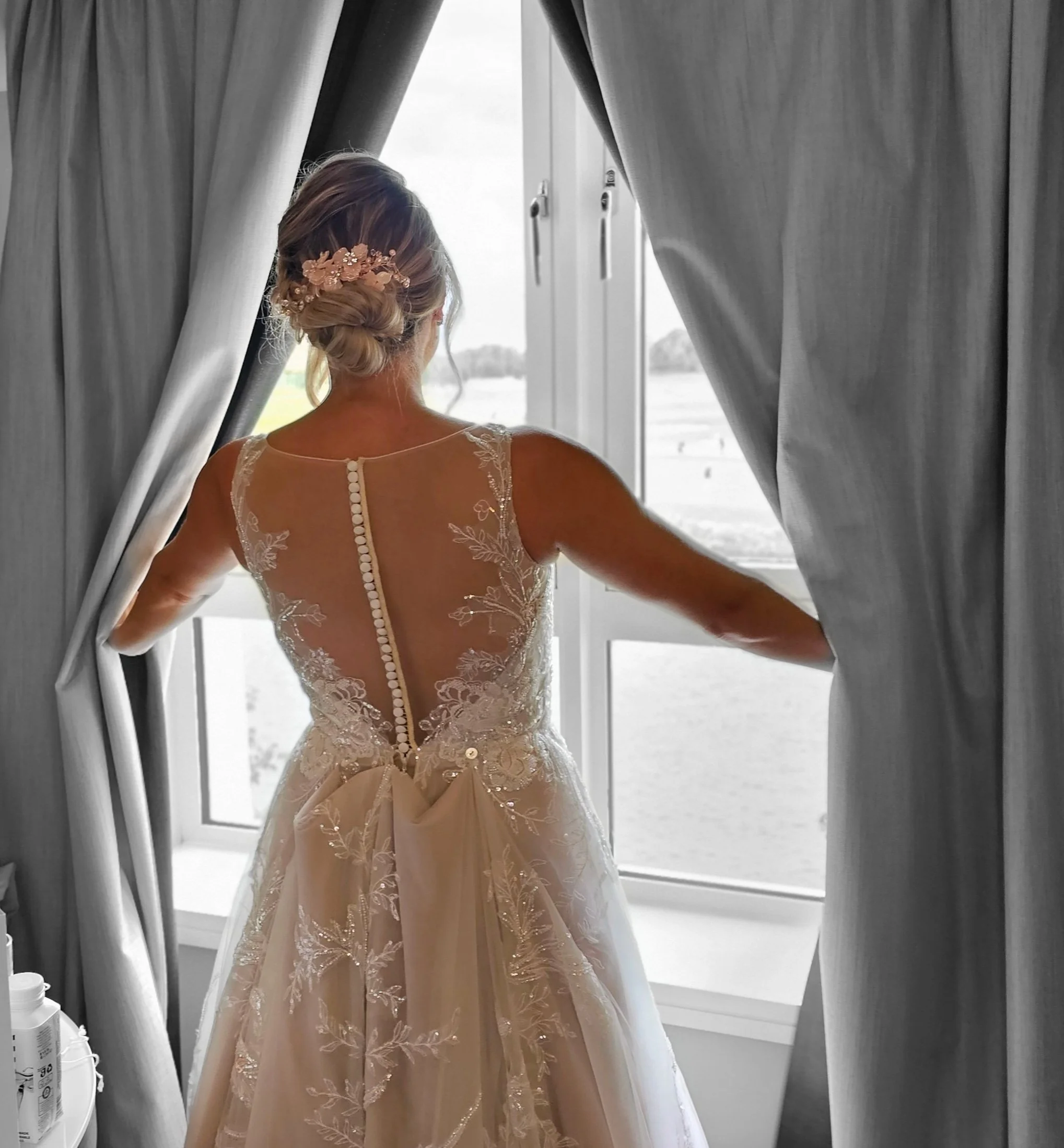 A woman in a wedding dress looking out of a window, with her back to the camera, adjusting the curtains. The dress features a sheer, embroidered back with button details.