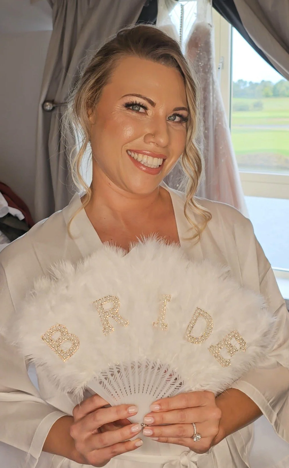 A smiling woman holding a white feather fan with the word 'BRIDE' spelled out in rhinestones, wearing a satin robe, standing indoors with a window showing a green landscape in the background.