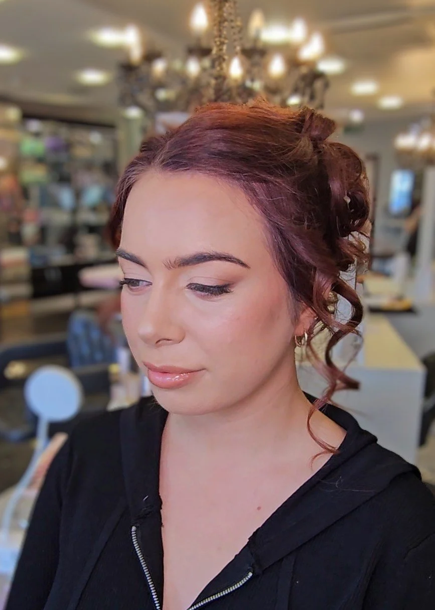 Close-up of a woman with red, curly hair styled in an elegant updo, wearing a black zip-up top, in a restaurant with chandeliers and blurred background.