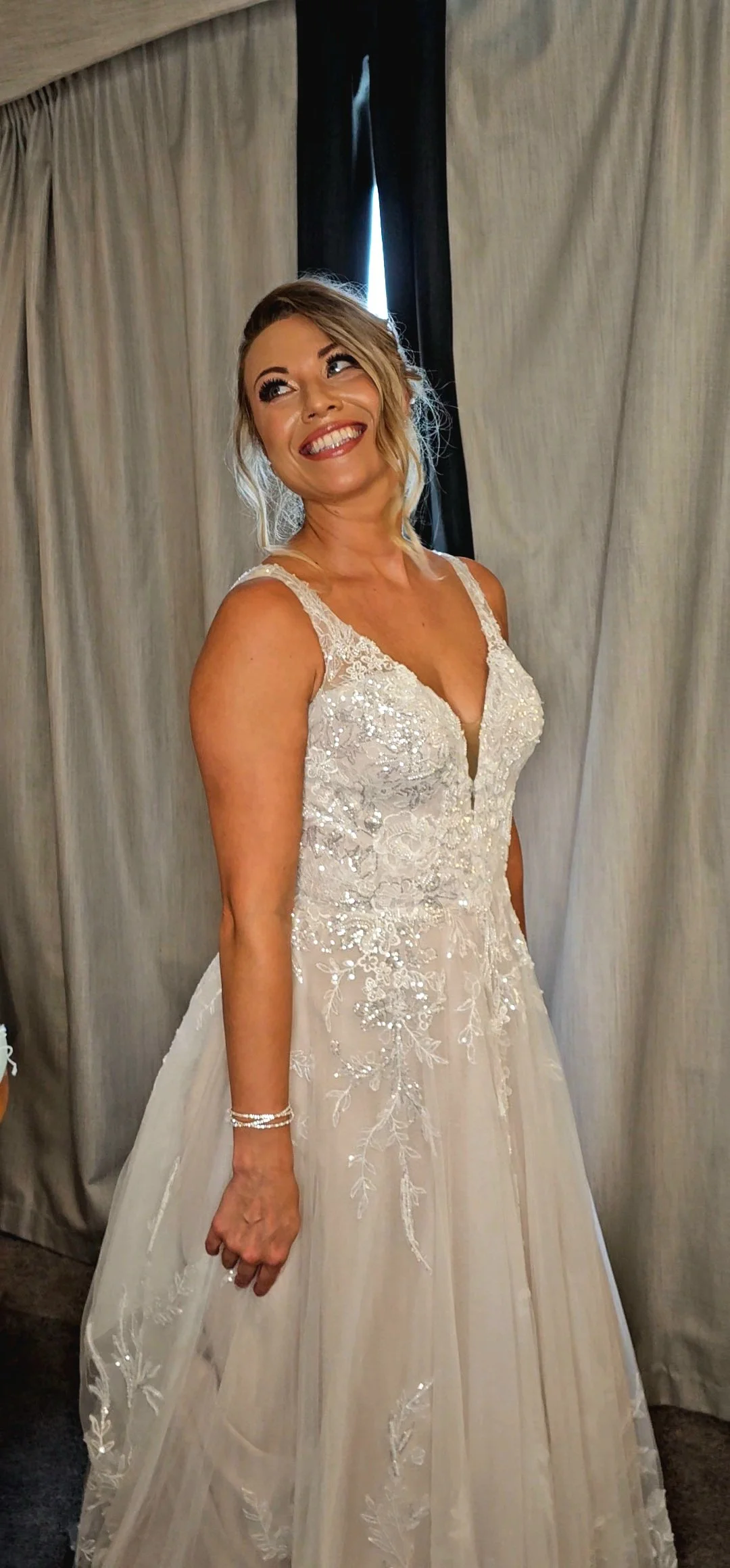 Woman in a white lace wedding dress smiling and looking to the side, standing in front of beige curtains with a dark opening behind her.