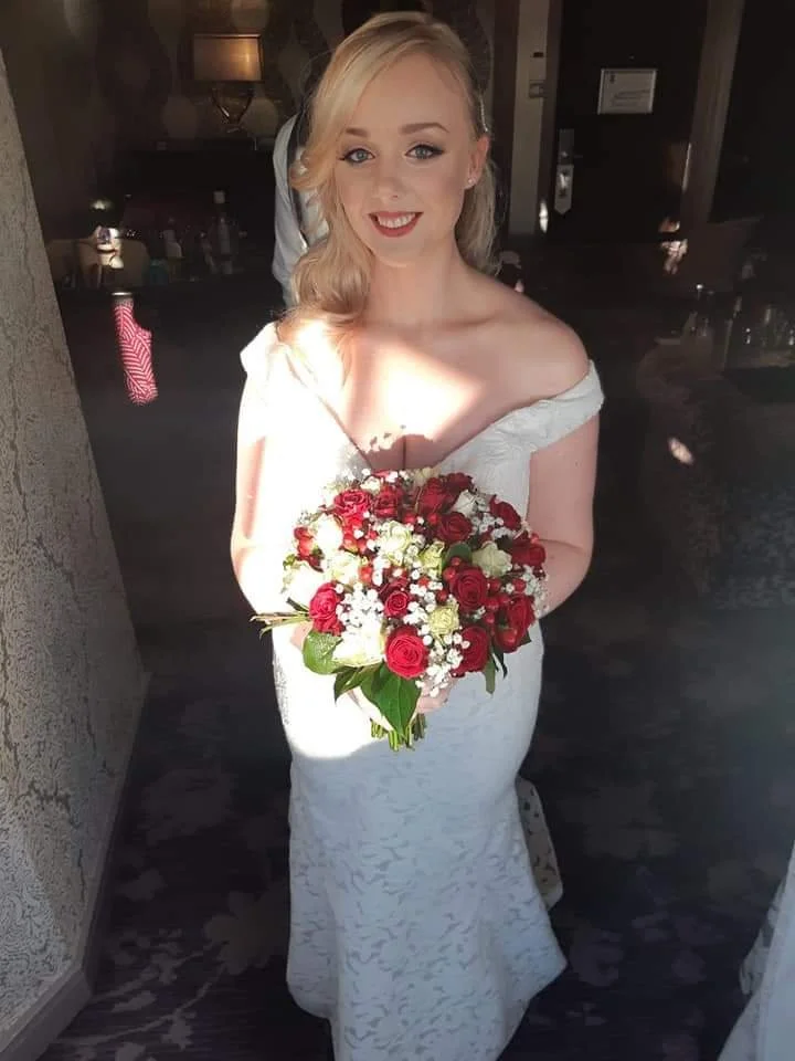 A bride in a white wedding dress holding a bouquet of red and white roses, smiling, with blonde hair styled in loose waves, standing indoors with sunlight on her face.