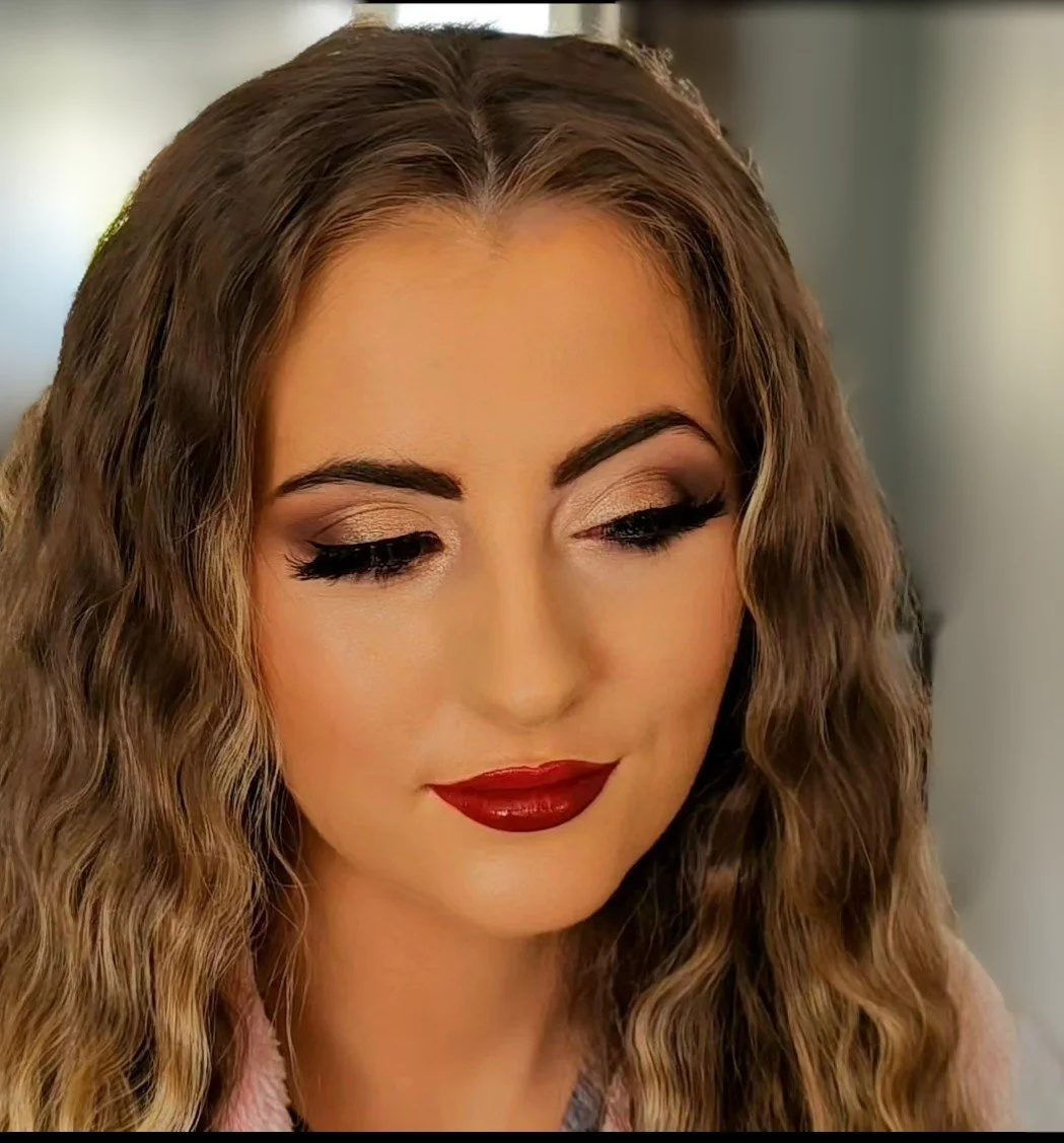 Close-up of a woman with long, wavy brown hair, makeup including gold eyeshadow, black eyeliner, mascara, and red lipstick, looking slightly downward.