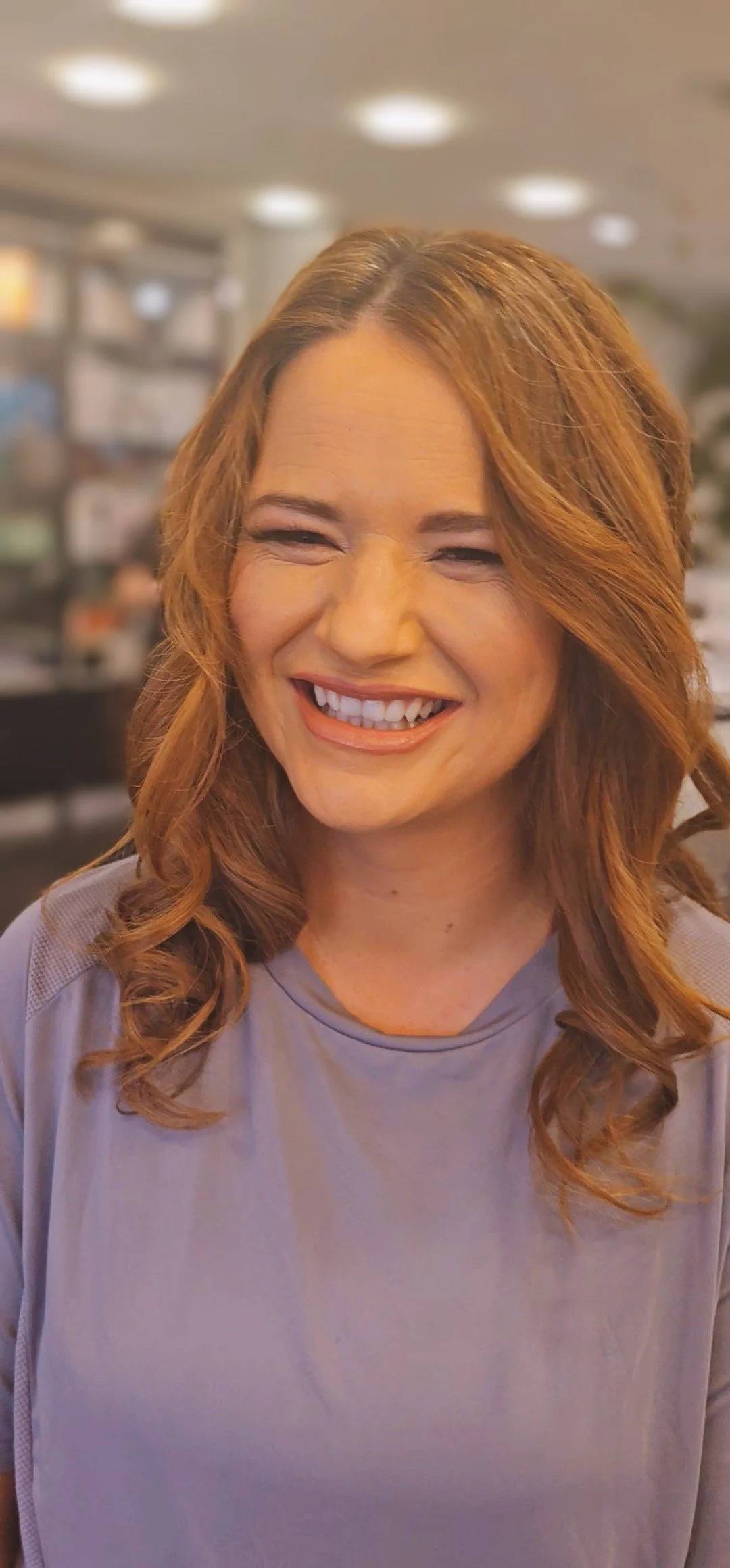 Close-up of a smiling woman with red hair and a gray top in an indoor setting, possibly a store or cafe.