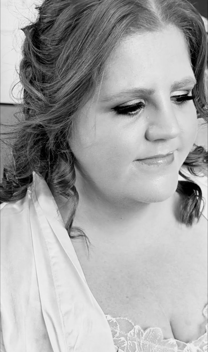 A black and white close-up portrait of a woman with wavy hair and makeup, looking down with a gentle expression.