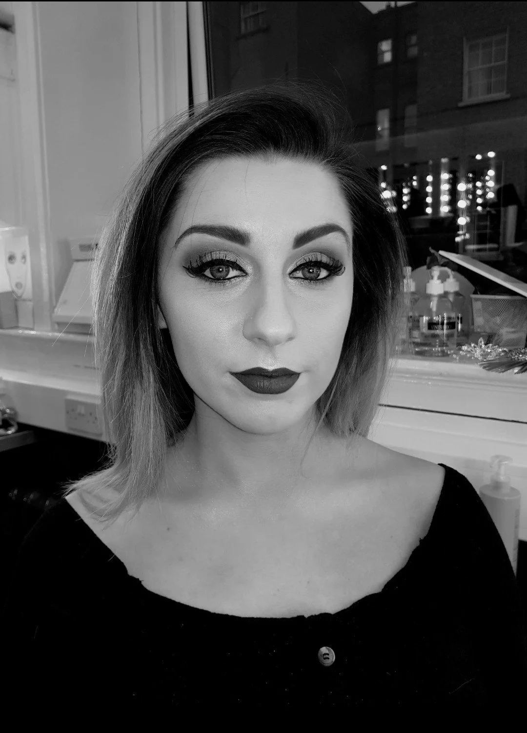Black and white portrait of a woman with makeup, dark lipstick, and shoulder-length hair, sitting indoors with a window and various items in the background.