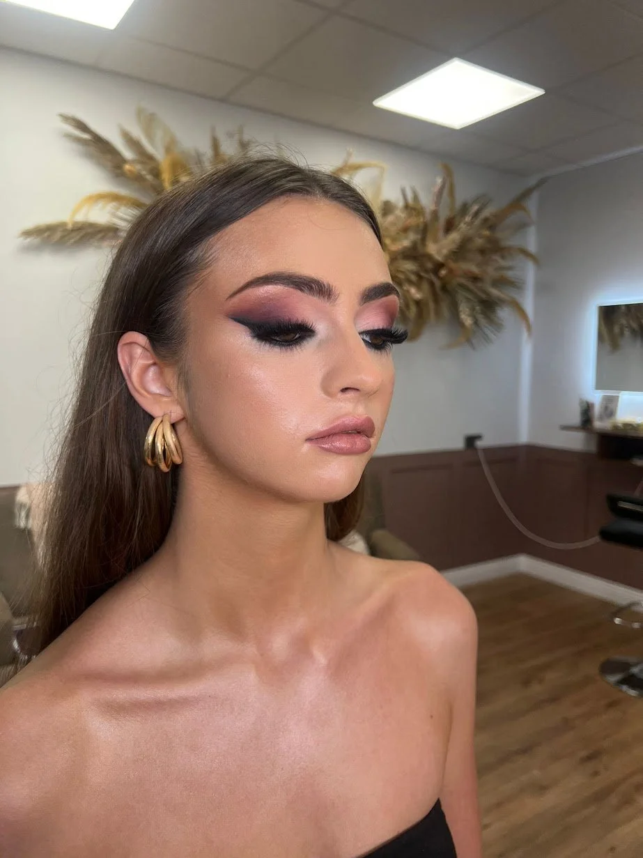 A woman with styled makeup, large gold hoop earrings, and long brown hair posing indoors.